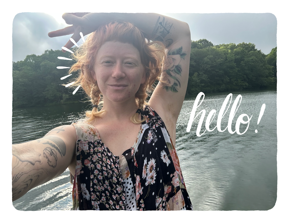 red haired woman with ivory skin and tattoos showing the peace sign in front of a river